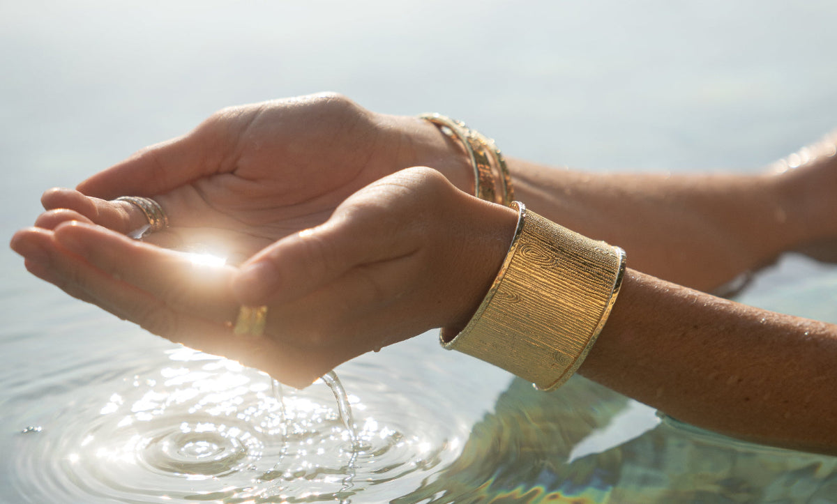 Hands with gold bracelets and a ring holding water, with a blurred natural background